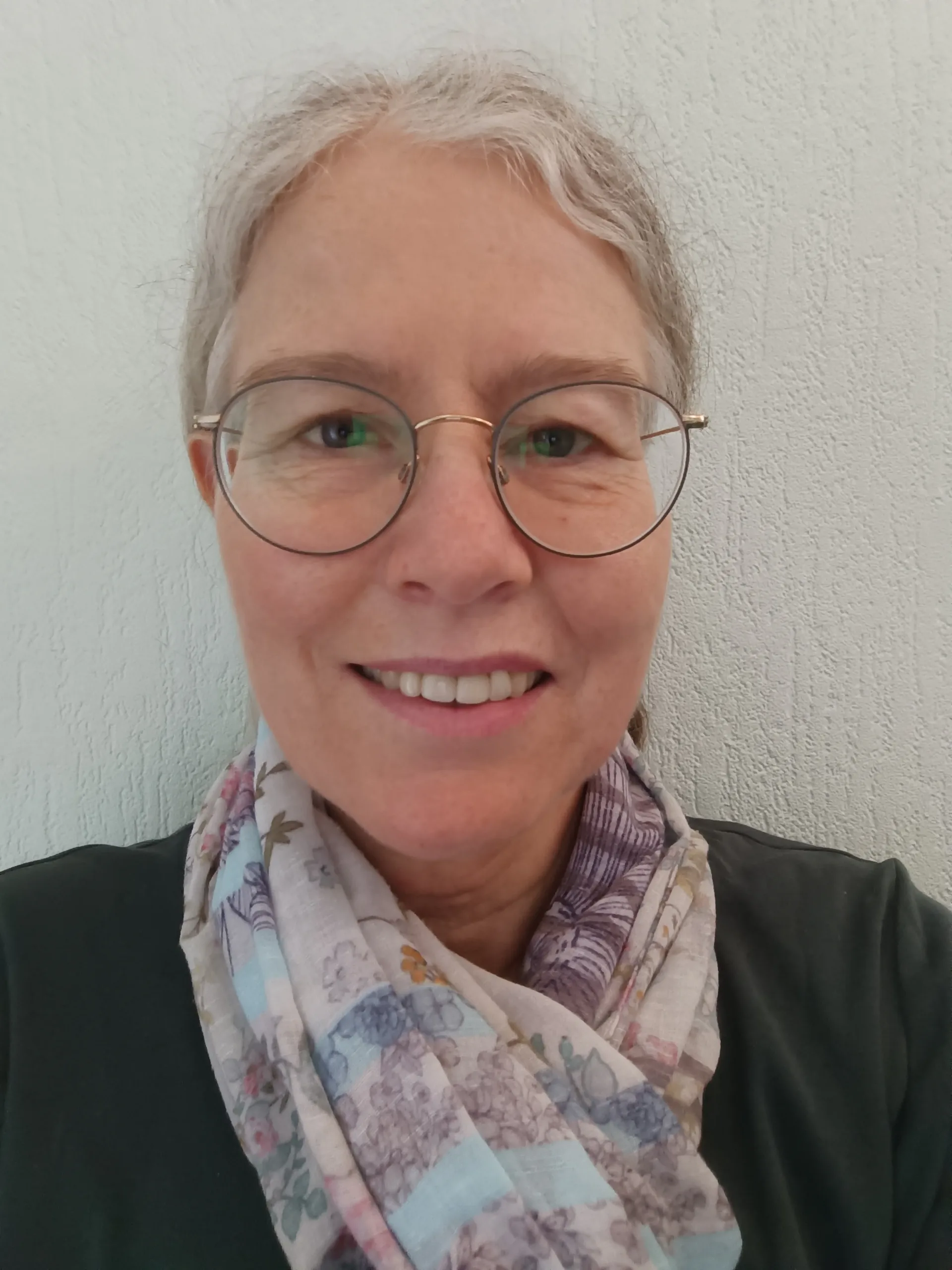 Abgebildet ist Petra im Portrait vor einer weißen Wand. Petra hat weiße Haare, die zu einem Zopf gebunden sind, und eine Brille. Sie trägt ein Halstuch mit Blumen und einen schwarzen Pullover. Ihr Gesicht ist lächelnd, aber leicht verzogen und angespannt.