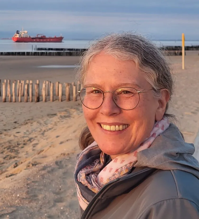 Zu sehen ist Petra im Portrait. Sie trägt eine Brille, hat zurückgebundene graue Harre und ein offenes Lächeln. Hinter ihr ist ein Strand sowie ein Frachtschiff zu sehen.