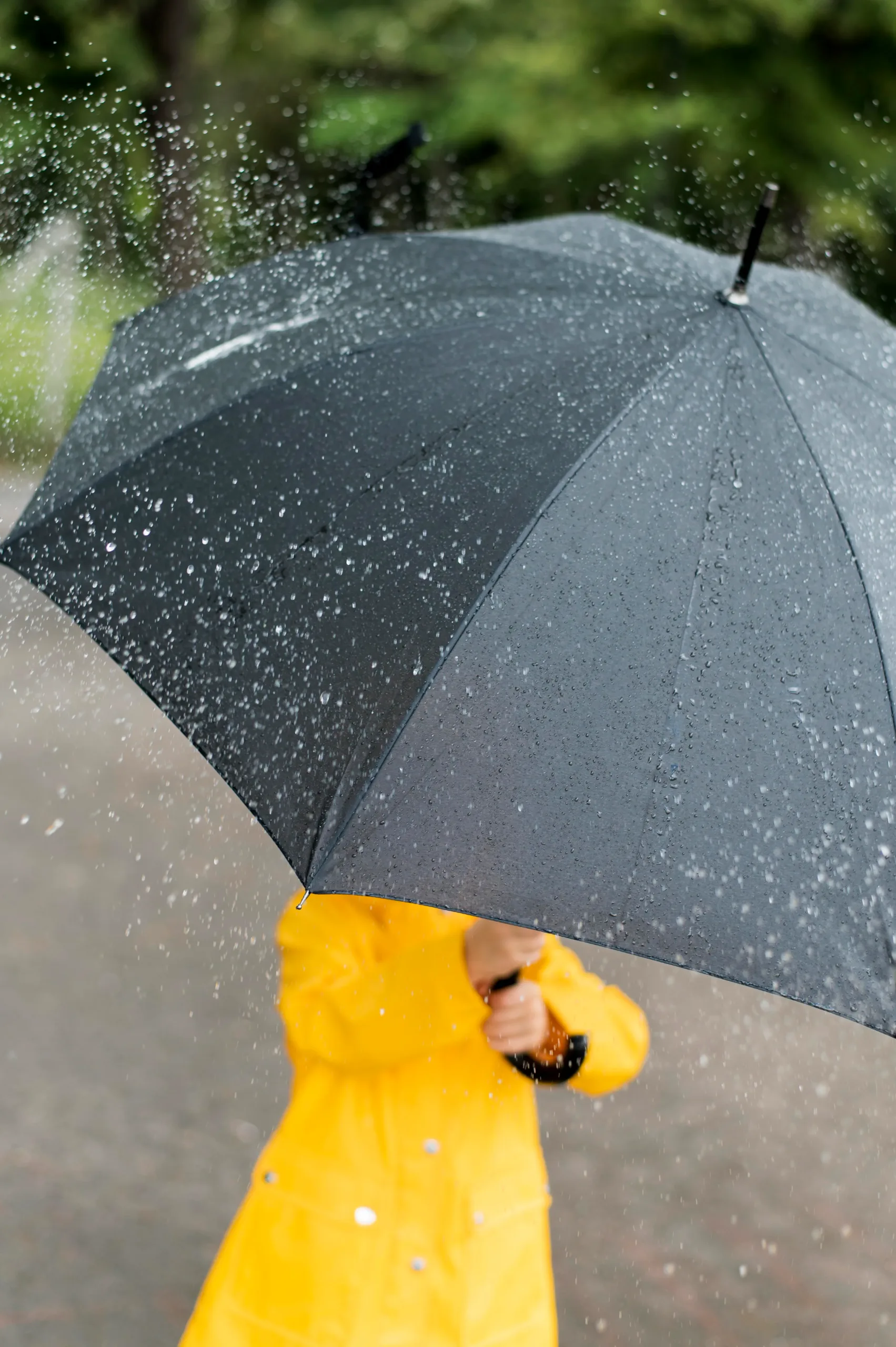 Person in gelbem Regenmantel, die einen großen schwarzen Schirm hält. Vom Schirm prallen Wassertropfen ab. Das Gesicht ist verdeckt.