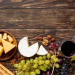 Zu sehen ist eine Auswahl von Käse, dazu ein Glas Rotwein und Trauben auf einer Holzunterlage